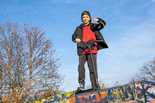 Ein netter und cooler Junge mit seinem schwarzen, trendigen Roller auf einem Spielplatz. Der Jugendliche ist sportbegeistert. Er fährt gern mit seinem Scooter oder Fahrrad (Bike).