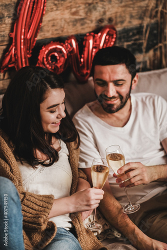 Young smiling happy couple in love in comfortable home clothing clinking glasses owith sparkling wine and celebrating holiday at home. Valentines day celebration concept photo