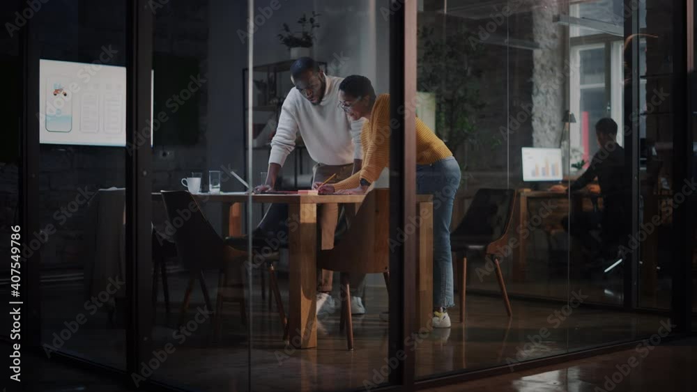 Two Diverse Multiethnic Colleagues Have a Conversation in a Meeting Room Behind Glass Walls in an Agency. African American Creative Director and Female Project Manager Discuss Work on Laptop Computer. Two Diverse Multiethnic Colleagues Have a Conversation in a Meeting Room Behind Glass Walls in an Agency. African American Creative Director and Female Project Manager Discuss Work on Laptop Computer.