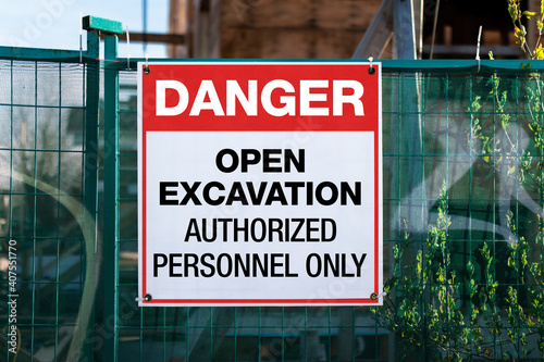 Construction site safety signage with text: Danger, open excavation, authorized personnel only. Large sign or poster on green construction fence. Defocused ply-wood building construction in progress.