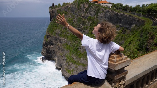 Back view of Caucasian woman sitting on an edge of observation deck with arms spread wide embracing unbelievable beauty of the nature photo