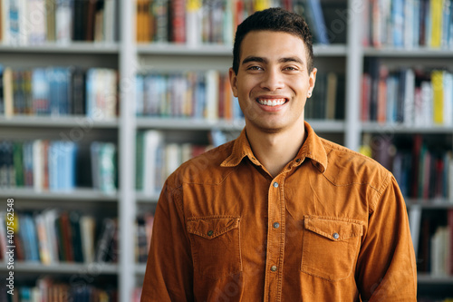 Portrait of successful hispanic business man looks directly at the camera, smile. Happy male freelancer or student standing in modern office or university library photo