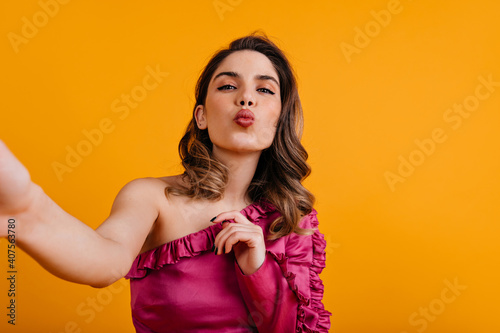 Glamorous curly woman posing with kissing face expression. Caucasian girl in silk blouse making selfie.