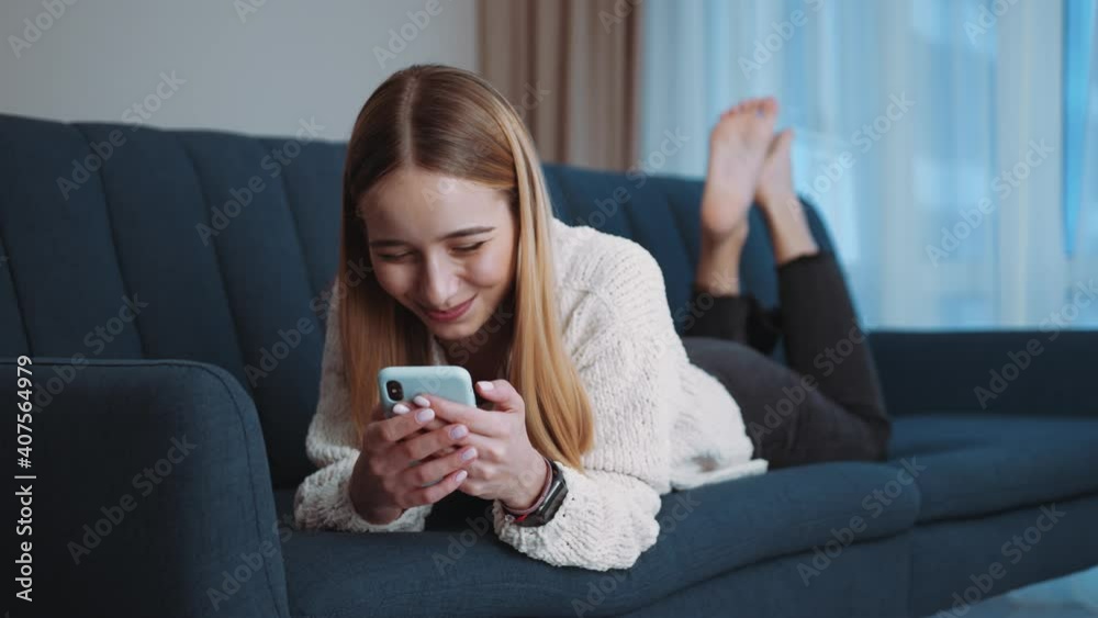 Happy young woman lying on sofa using smartphone in modern room. Smiling. Social media. Relax. Tapping, scrolling. Internet online connection. Slow motion Happy young woman lying on sofa using smartphone in modern room. Smiling. Social media. Relax. Tapping, scrolling. Internet online connection. Slow motion