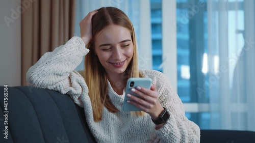 Cute young woman sitting on sofa using smartphone in modern room. Happy, smiling. Social media. Relax. Tapping, scrolling. Internet online connection. Slow motion