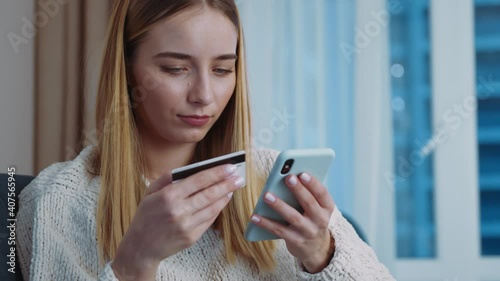 Young woman holding credit card using mobile payments sitting on sofa at home. Making purchase on smart phone receiving cashback. Finance credit banking money transaction. Slow motion