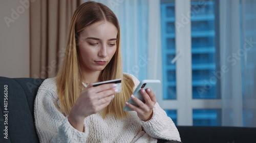 Smiling woman holding credit card using mobile payments sitting on sofa at home. Making purchase on smart phone receiving cashback. Finance credit banking money transaction. Slow motion