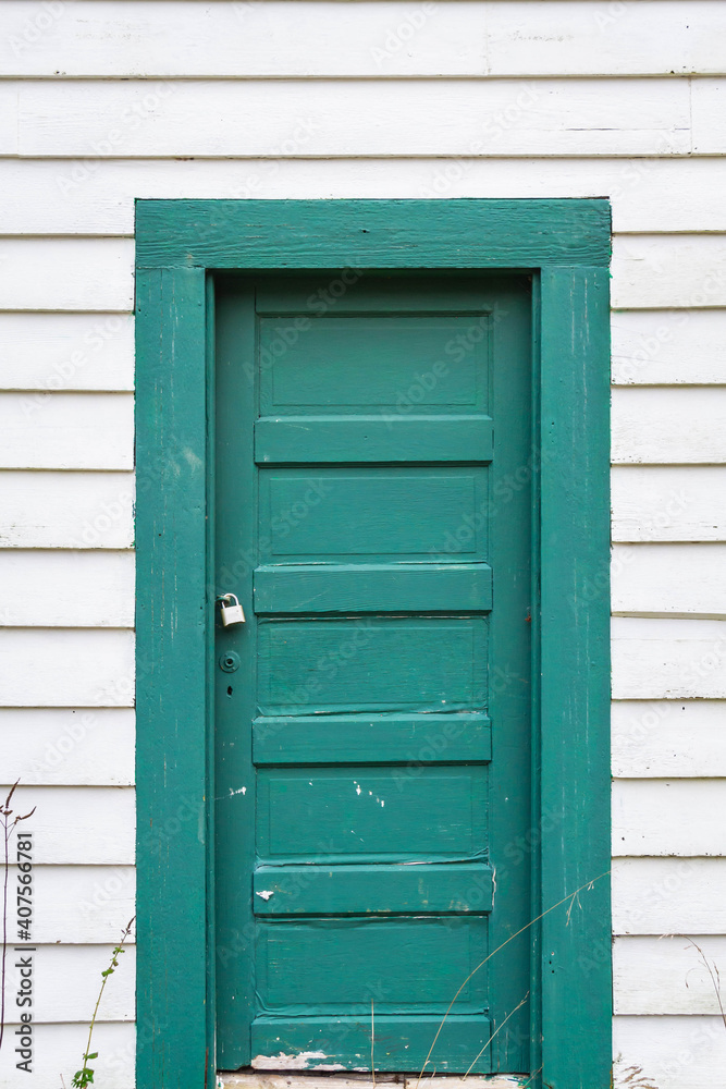 An old green door on the side of a white building with some weeds on the bottom of the image.