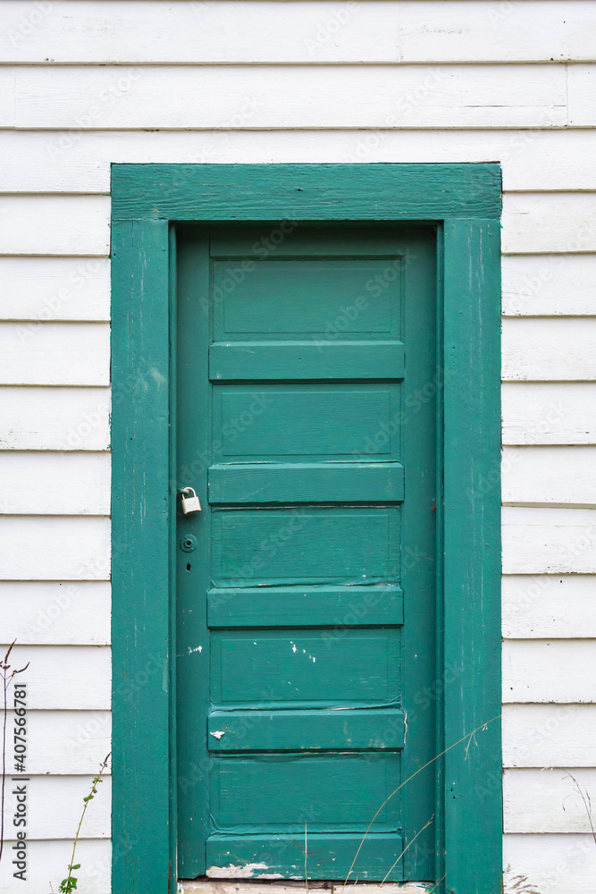 An old green door on the side of a white building with some weeds on the bottom of the image. An old green door on the side of a white building with some weeds on the bottom of the image.