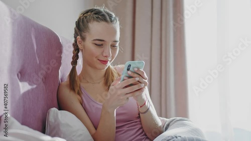 At sunlight attractive woman holding mobile phone while lying in bed at home. Morning time. Social media. Communication telephone texting bedroom. Slow motion