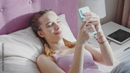 Smiling attractive woman holding mobile phone while lying in bed at sunlight in home. Morning time. Social media. Communication telephone texting bedroom. Slow motion