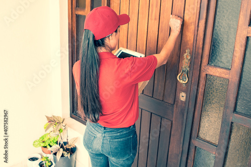 Latin courier knocking on door, holding tablet and carton boxes. Brunette long-haired deliverywoman in red uniform standing in front of door and delivering order. Delivery service and post concept photo