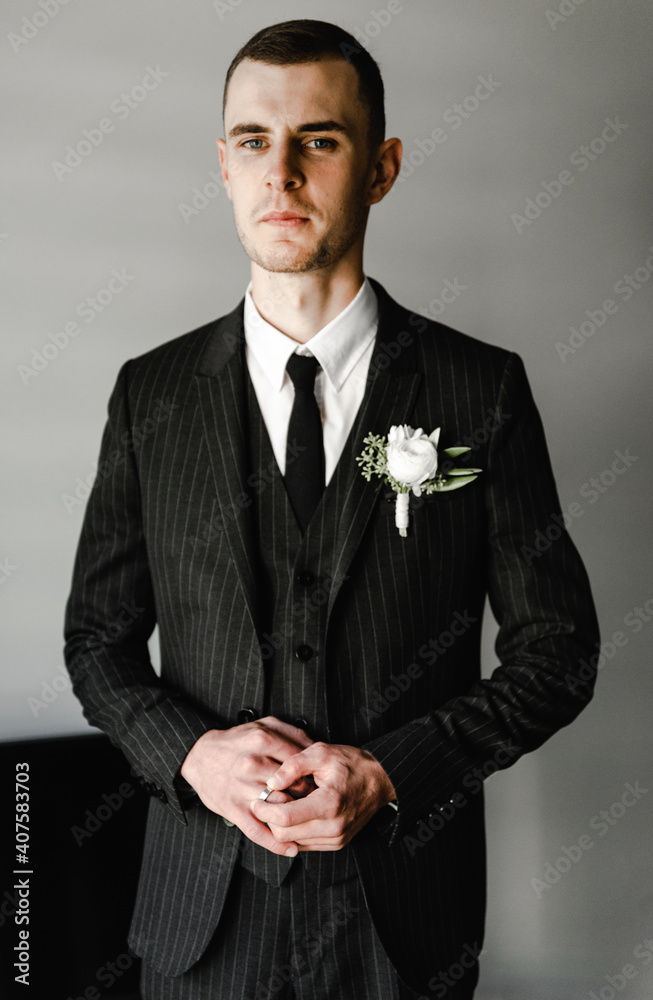 groom in a black suit stands against the gray background of his room. fees of the groom. wedding day. the groom is nervous before the wedding ceremony groom in a black suit stands against the gray background of his room. fees of the groom. wedding day. the groom is nervous before the wedding ceremony