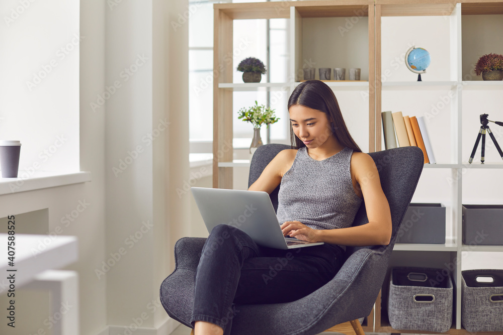 Young positive asian woman office worker sitting in armchair with laptop and working online Young positive asian woman office worker sitting in armchair with laptop and working online