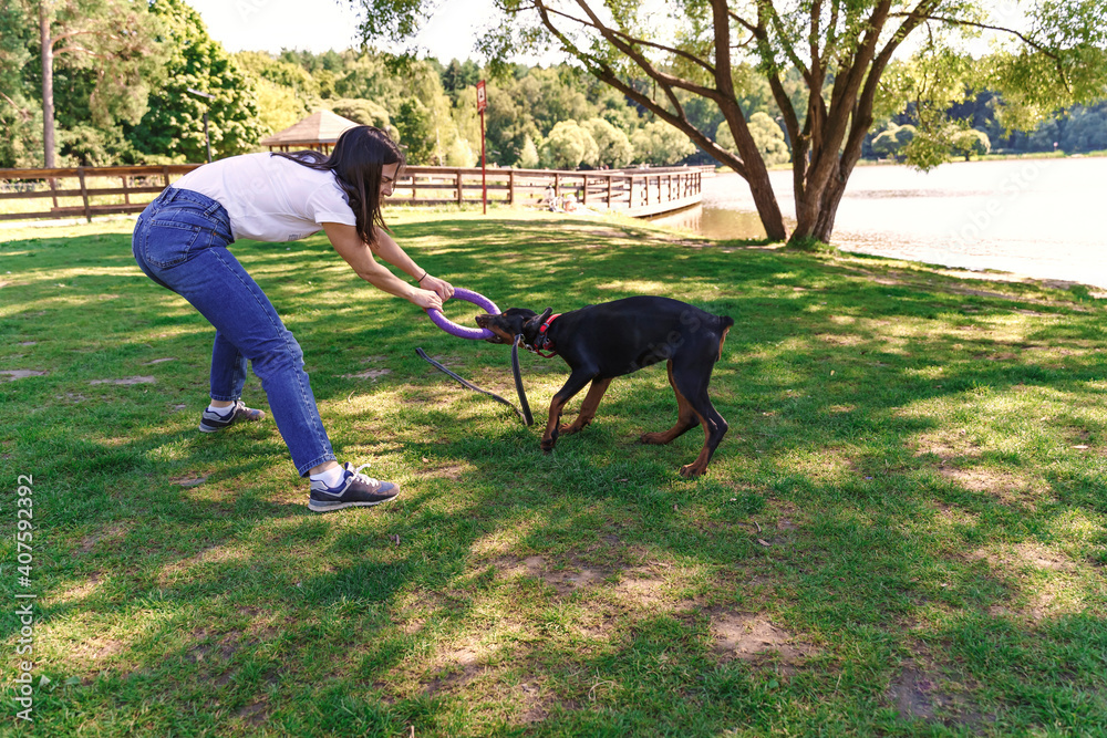 Cheerful brunette woman spends time with soy dog Doberman by the lake. puppy playing with a toy ring. Walking dogs in nature. Cheerful brunette woman spends time with soy dog Doberman by the lake. puppy playing with a toy ring. Walking dogs in nature.