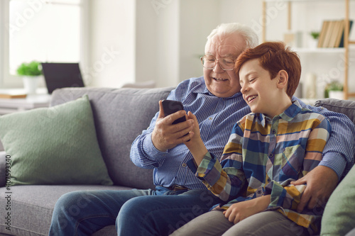 Smiling grandfather and grandson sitting on sofa and having fun. Little child showing grandpa how to use social media app on smartphone. Happy elderly man learning to make video call on mobile phone photo