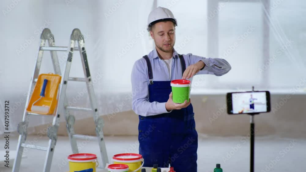 repairs blog, young male professional builder in helmet talks about building paints for walls and points to camera on smartphone during live indoor broadcast repairs blog, young male professional builder in helmet talks about building paints for walls and points to camera on smartphone during live indoor broadcast