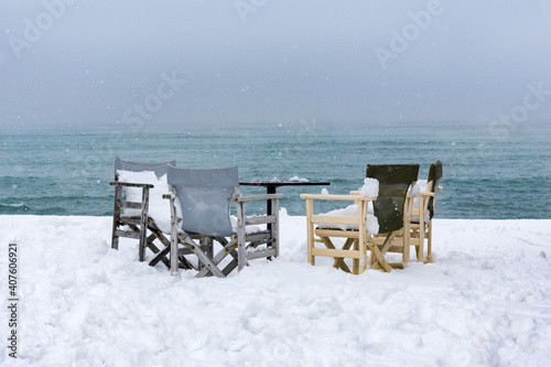 Four director's chairs and a table full of snow, on the edge of the snowy pier against the backdrop of the rough sea