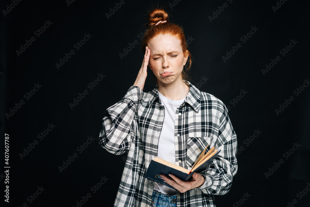 Naklejka premium Crying upset young woman college student with headache holding book and touching temples on isolated black background. Pretty redhead lady model emotionally showing facial expressions in studio