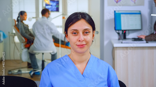 Dental assistant looking at camera while doctor examining patient in background. Professional stomatologist nurse smiling on webcam sitting on chair in waiting room of stomatological clinic.