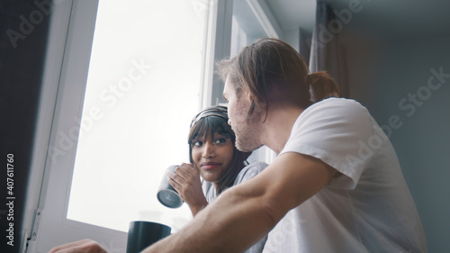 Young happy multiracial couple drinking coffee or tea and looking through the window. Slow motion. High quality photo