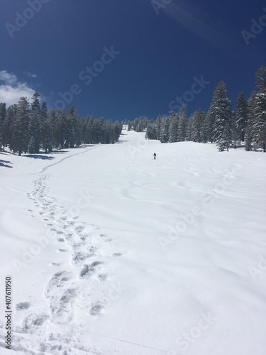 A snowboard hikes up a mountain with poles and board on his back, leaving boot tracks in the snow