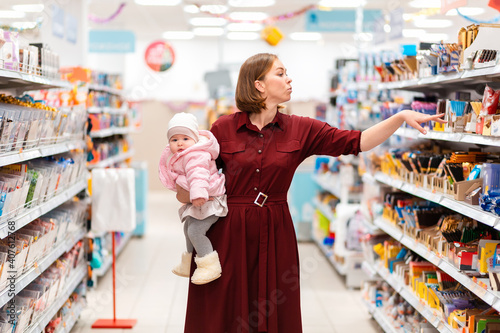 Shopping and consumerism. Portrait of a young mother holding her baby in her arms and choosing products on a supermarket shelf. The concept of family shopping