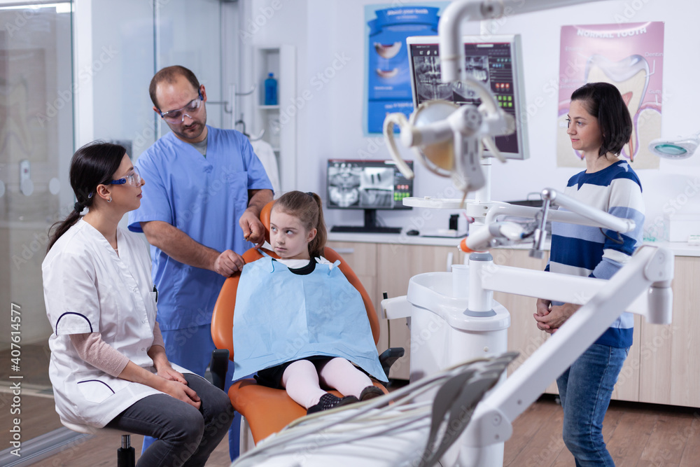 Kid patient in dentist office prepared for stomatology treatment while parent is talking with doctor. Child with her mother during teeth check up with stomatolog sitting on chair. Kid patient in dentist office prepared for stomatology treatment while parent is talking with doctor. Child with her mother during teeth check up with stomatolog sitting on chair.