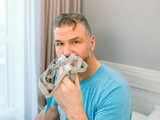 Unhappy shocked man with chronic breathing issues surprised by using  CPAP machine sitting on the bed in bedroom. Healthcare, CPAP, Obstructive sleep apnea therapy, snoring concept