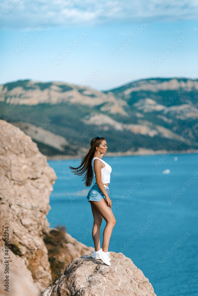 Joyful woman travel mountains. Authentic blond female relax in picturesque landscape. A girl with developing natural hair is wearing a white top and denim shorts.