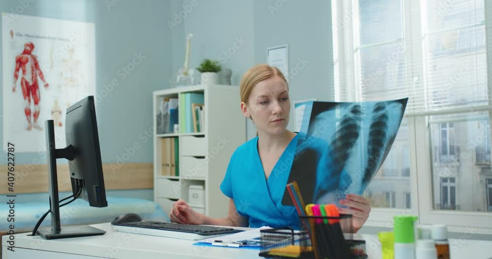 Pretty young hopital worker looking at x-ray scan while sitting at table. Professional Caucasian female nurse in blue medical gown documenting report on disease. Pneumonia, medicine, healthcare.