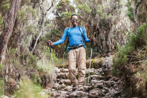 Portrait of a cheerfully smiling African-American Ethnicity young man in sunglasses. He having a walk with a backpack using trekking poles to Mweka gate, Kilimanjaro. Active climbing people concept.