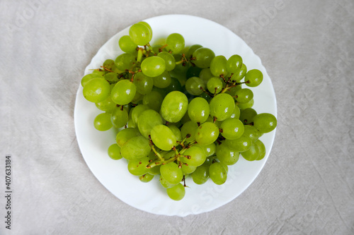 Grona of white seedless grapes in a white plate on a beige linen tablecloth top view . raw fruit on a plate