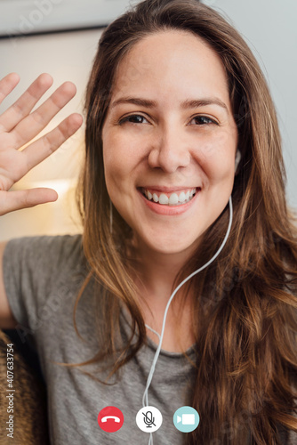 Happy young woman waving on video call meeting with mobile phone inside home during lockdown isolation - Technology, social distance communication concept -Focus on face photo