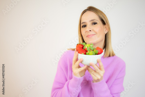 Beautiful cheerful young woman standing and holding glass bowl with strawberries over white background