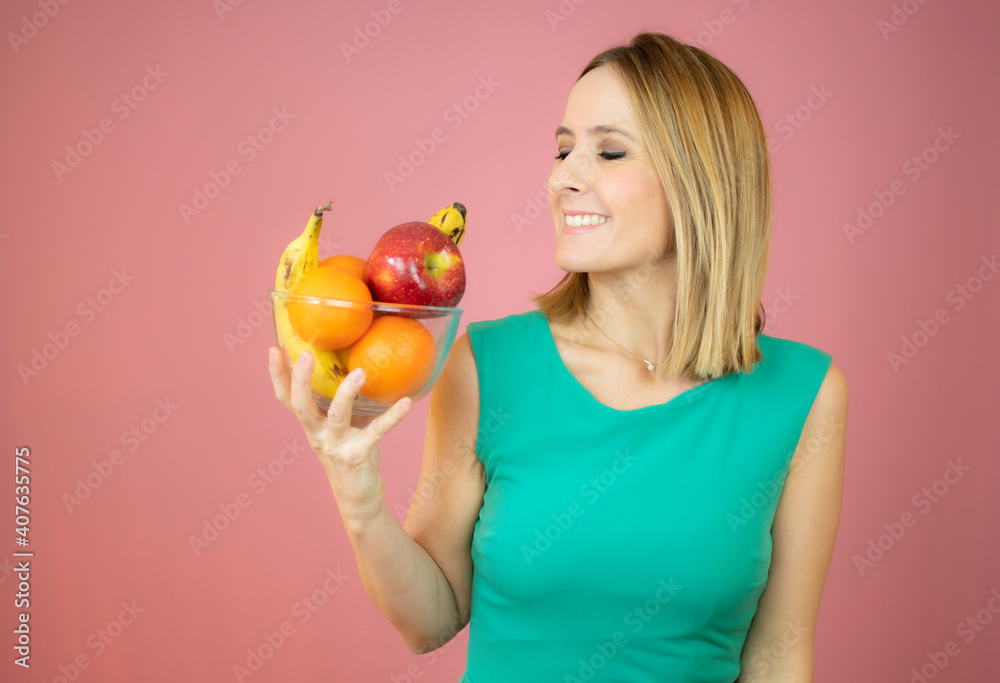 Beautiful cheerful young woman standing and holding glass bowl with fruits over pink background Beautiful cheerful young woman standing and holding glass bowl with fruits over pink background