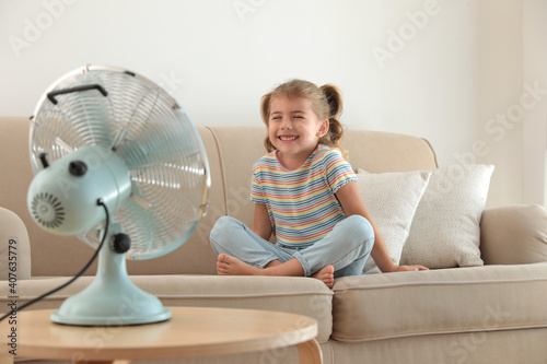Little girl enjoying air flow from fan on sofa in living room. Summer heat photo