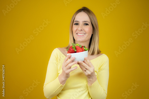 Beautiful cheerful young woman standing and holding glass bowl with strawberries over yellow background