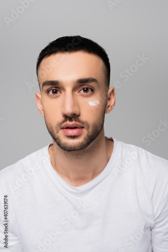 young hispanic man with stroke of cosmetic cream on face looking at camera isolated on grey