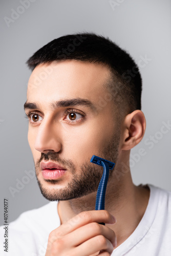 bearded hispanic man shaving with safety razor isolated on grey