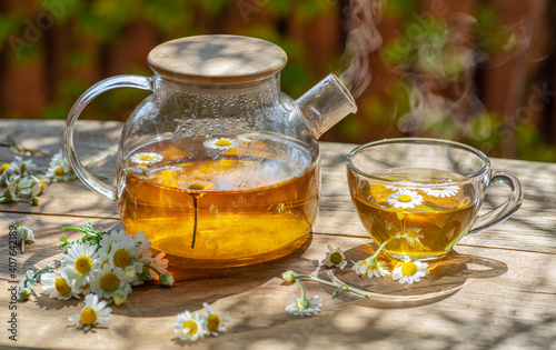 Herbal chamomile tea and chamomile flowers near teapot and tea glass on wooden table. Countryside background.