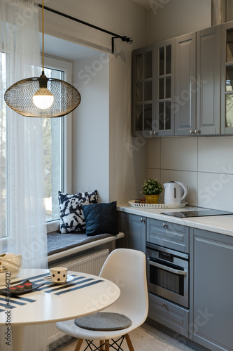 Interior of the newly equipped kitchen part in the studio apartment. Round table with lamp  chair by the window and wide windowsill with cushions. Kitchen furniture with oven  hob  beige countertop