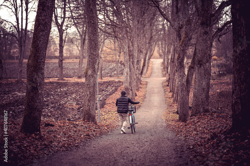 Person walking with his bike in a small path in forest between big trees a grey and cold autumn day