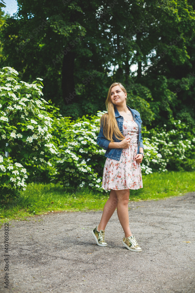 A young woman of European appearance with long blond hair, dressed in a short dress, stands against the background of white flowering bushes. Sunny spring day. Natural female beauty A young woman of European appearance with long blond hair, dressed in a short dress, stands against the background of white flowering bushes. Sunny spring day. Natural female beauty