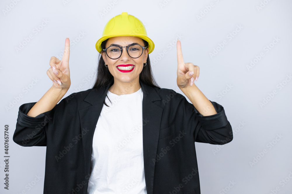Young architect woman wearing hardhat smiling, looking at the camera and pointing up with fingers and raised arms Young architect woman wearing hardhat smiling, looking at the camera and pointing up with fingers and raised arms