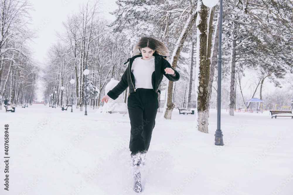 A beautiful young girl in warm clothes walks, plays and poses against the background of a winter snow-covered park and a playground