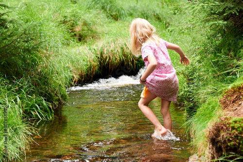 Child cute blond girl playing in the creek. Gril walking in forest stream and exploring nature. Summer children fun. Children summer activities