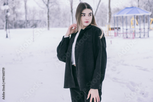 A beautiful young girl in warm clothes walks, plays and poses against the background of a winter snow-covered park and a playground