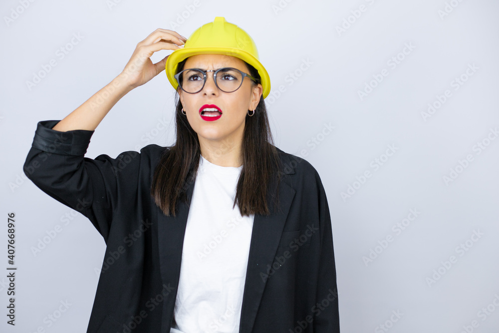 Young architect woman wearing hardhat confuse and wonder about question. Uncertain with doubt, thinking with hand on head