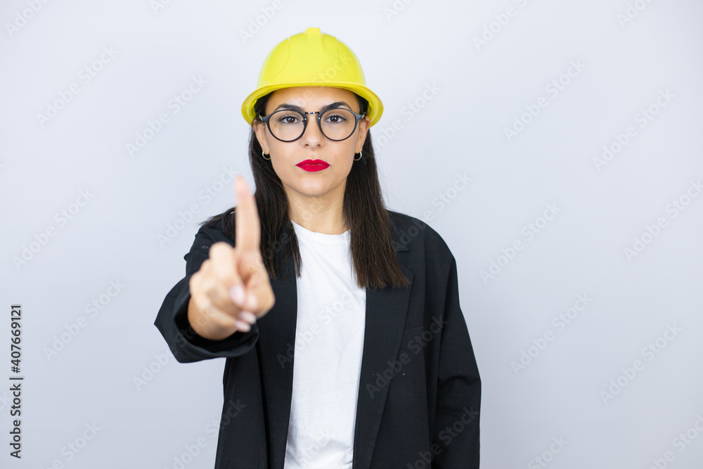 Young architect woman wearing hardhat showing and pointing up with fingers number one while is serious